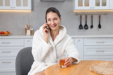 Photo of Beautiful woman with glass of juice talking on smartphone after spa procedure in kitchen