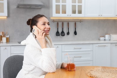 Photo of Beautiful woman with glass of juice talking on smartphone after spa procedure in kitchen