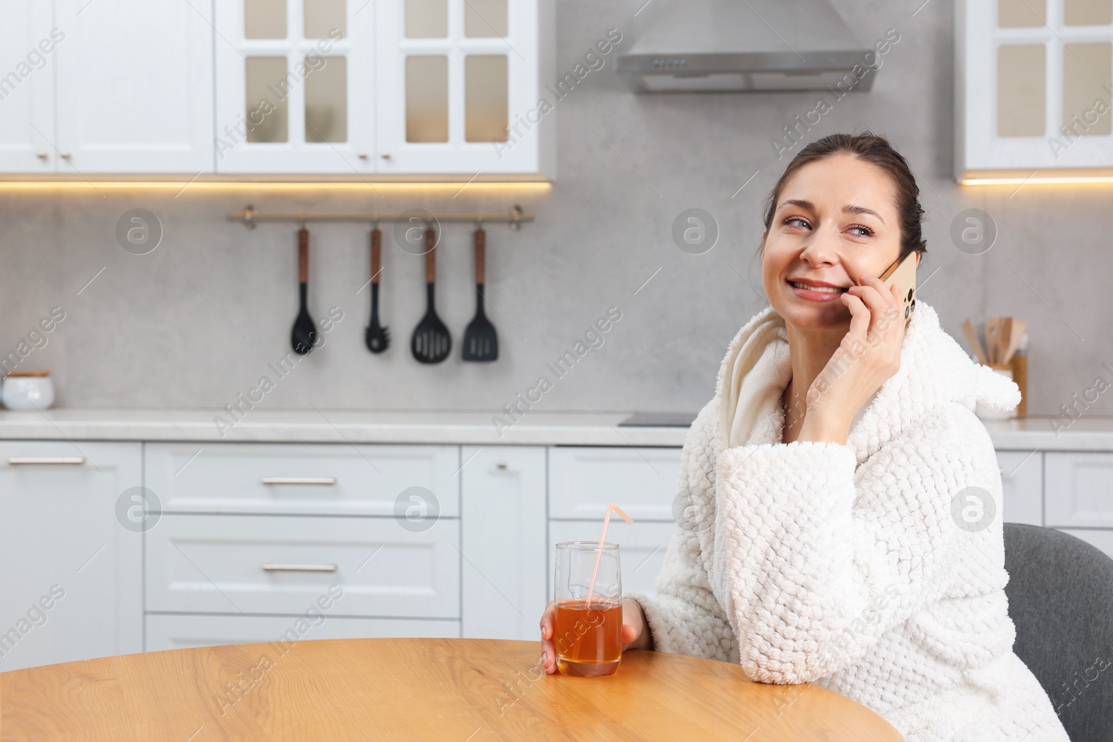 Photo of Beautiful woman with glass of juice talking on smartphone after spa procedure in kitchen