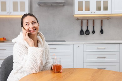 Photo of Beautiful woman with glass of juice talking on smartphone after spa procedure in kitchen