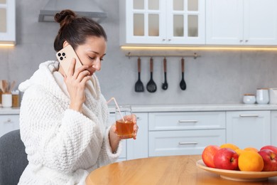 Photo of Beautiful woman with glass of juice talking on smartphone after spa procedure in kitchen