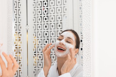 Photo of Spa day. Beautiful woman applying mask onto her face near mirror indoors