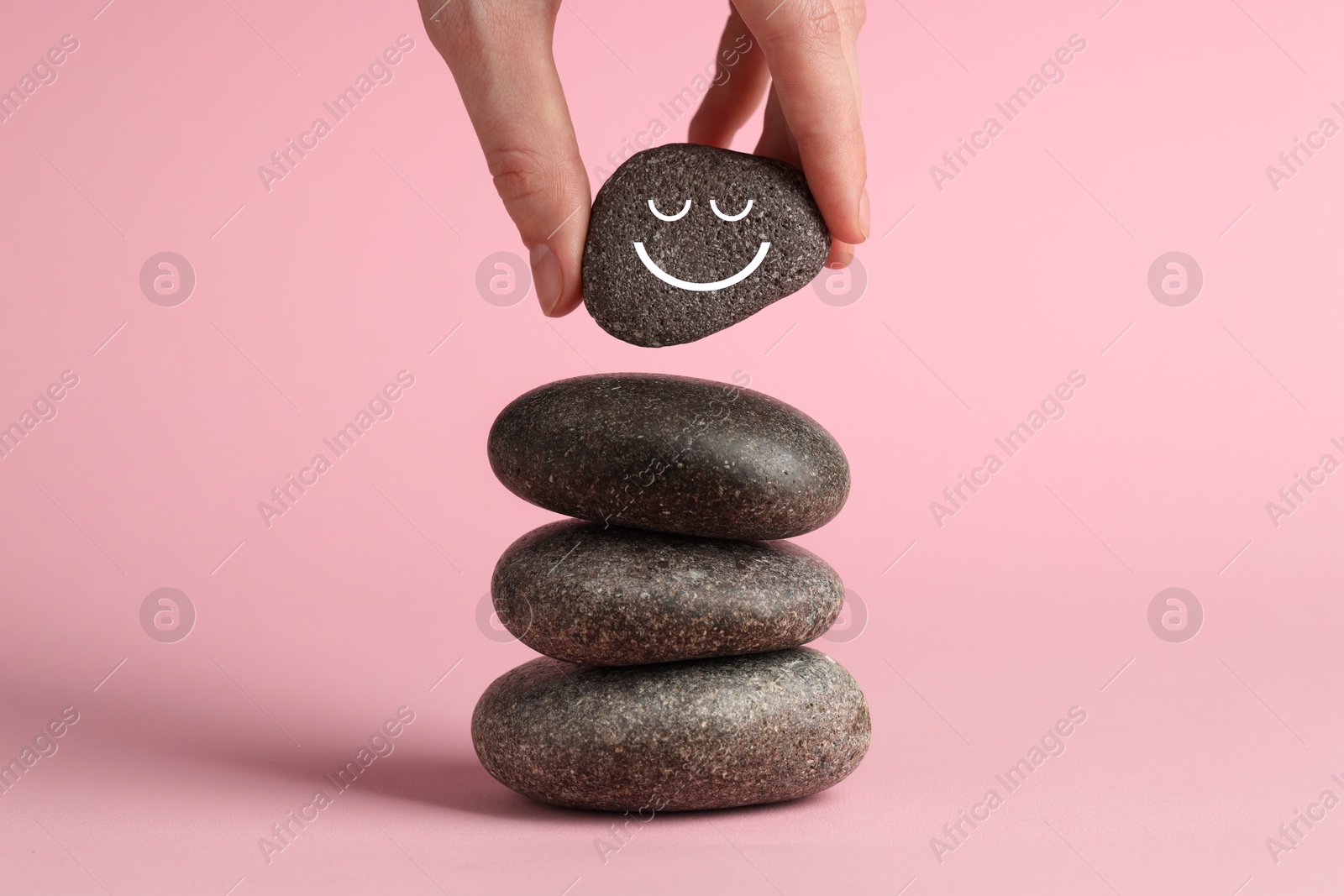 Photo of Woman putting rock with smiley face onto cairn against pink background, closeup. Harmony and life balance