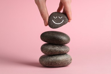 Photo of Woman putting rock with smiley face onto cairn against pink background, closeup. Harmony and life balance