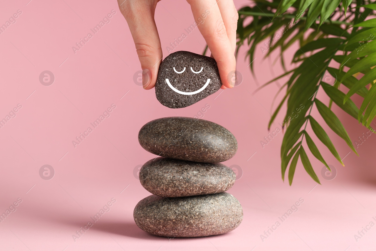 Photo of Woman putting rock with smiley face onto cairn against pink background, closeup. Harmony and life balance