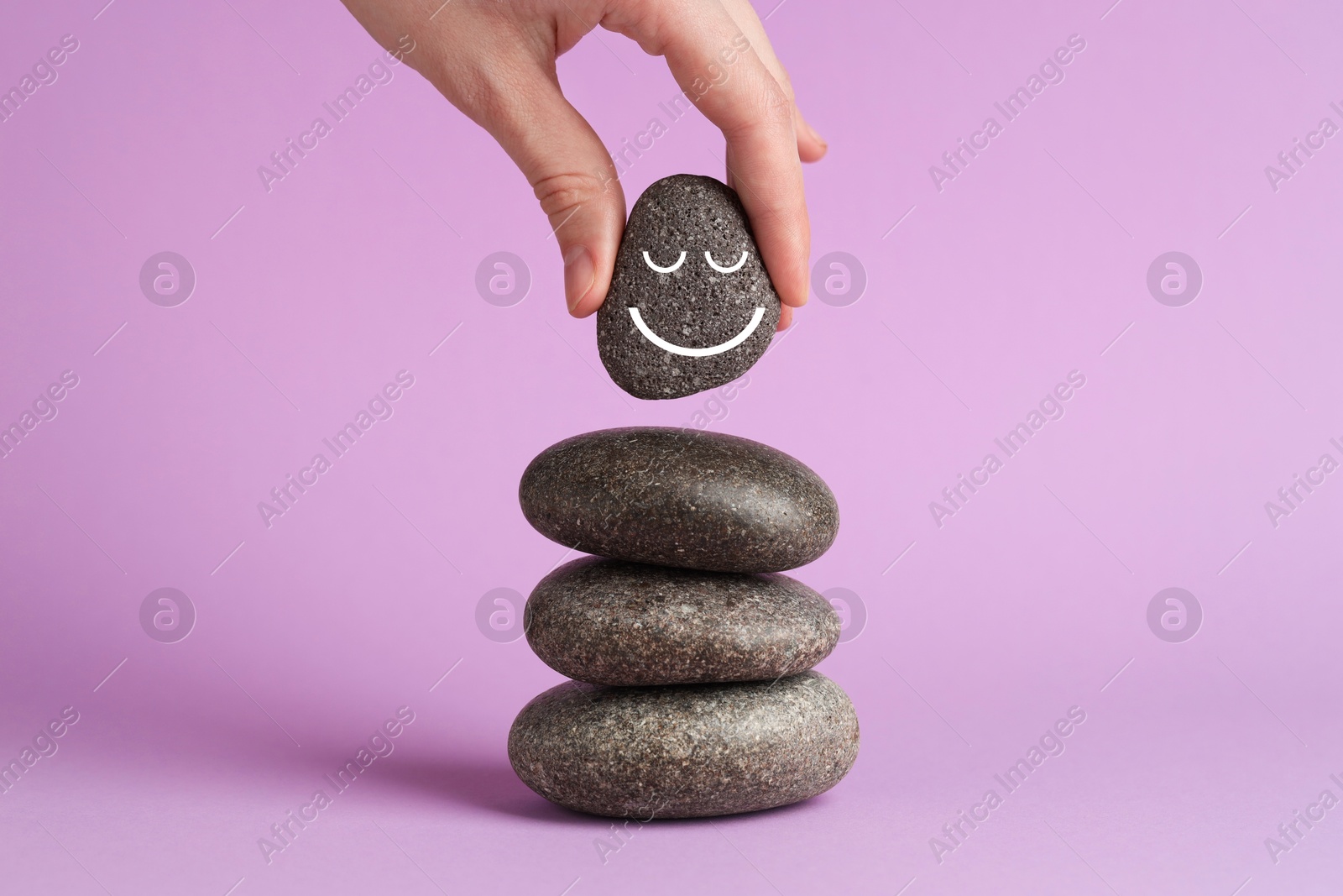 Photo of Woman putting rock with smiley face onto cairn against lilac background, closeup. Harmony and life balance