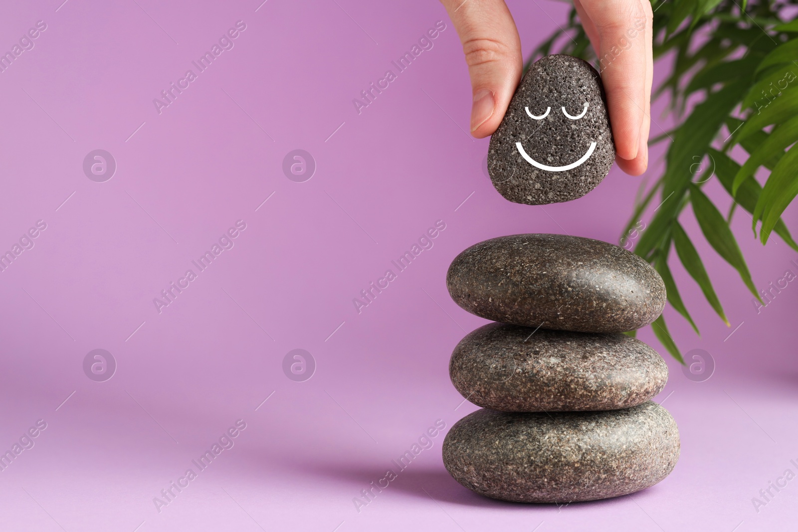 Photo of Harmony and life balance. Woman putting rock with smiley face onto cairn against lilac background, closeup. Space for text