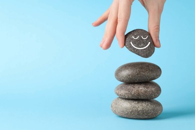 Photo of Harmony and life balance. Woman putting rock with smiley face onto cairn against light blue background, closeup. Space for text