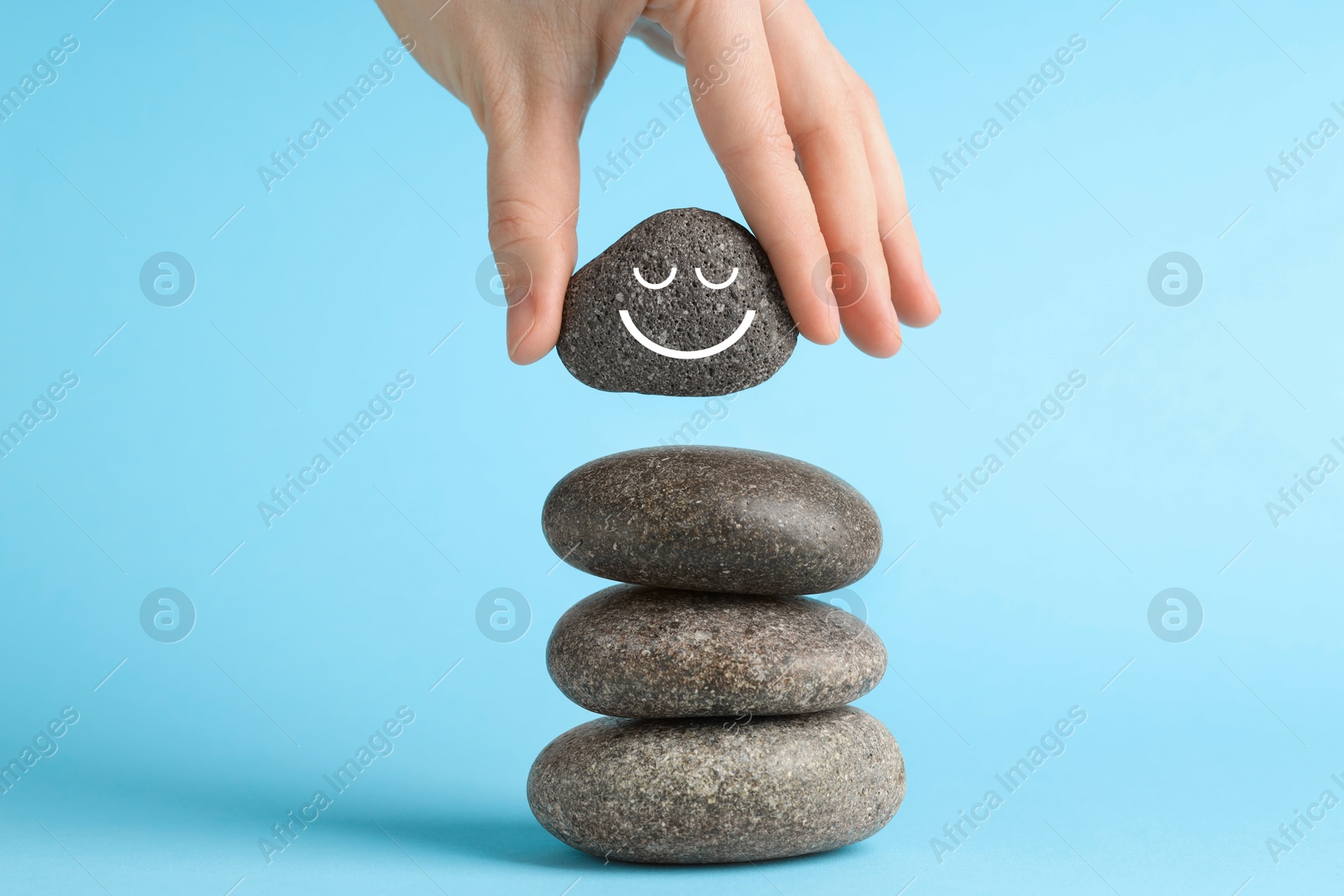 Photo of Woman putting rock with smiley face onto cairn against light blue background, closeup. Harmony and life balance