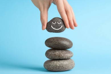 Photo of Woman putting rock with smiley face onto cairn against light blue background, closeup. Harmony and life balance