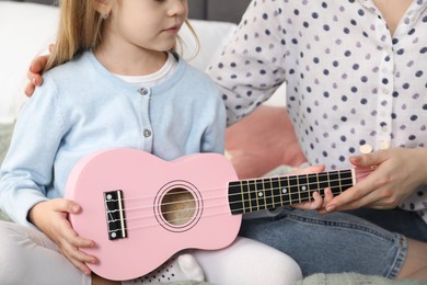 Photo of Woman teaching little girl to play ukulele at home, closeup