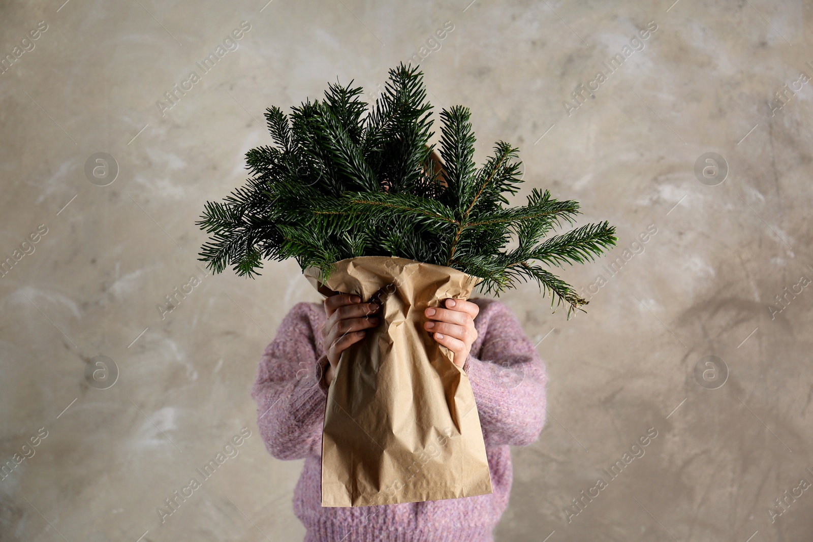 Photo of Woman holding green fir branches in paper bag near gray wall. Christmas decor