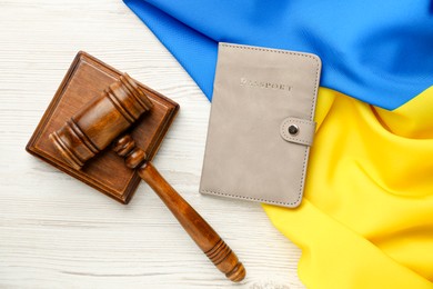 Photo of Passport in leather cover, gavel and flag of Ukraine on light wooden table, top view