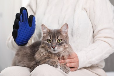 Photo of Woman brushing her cat with grooming glove indoors, closeup