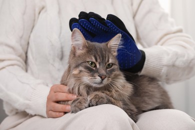 Photo of Woman brushing her cat with grooming glove indoors, closeup