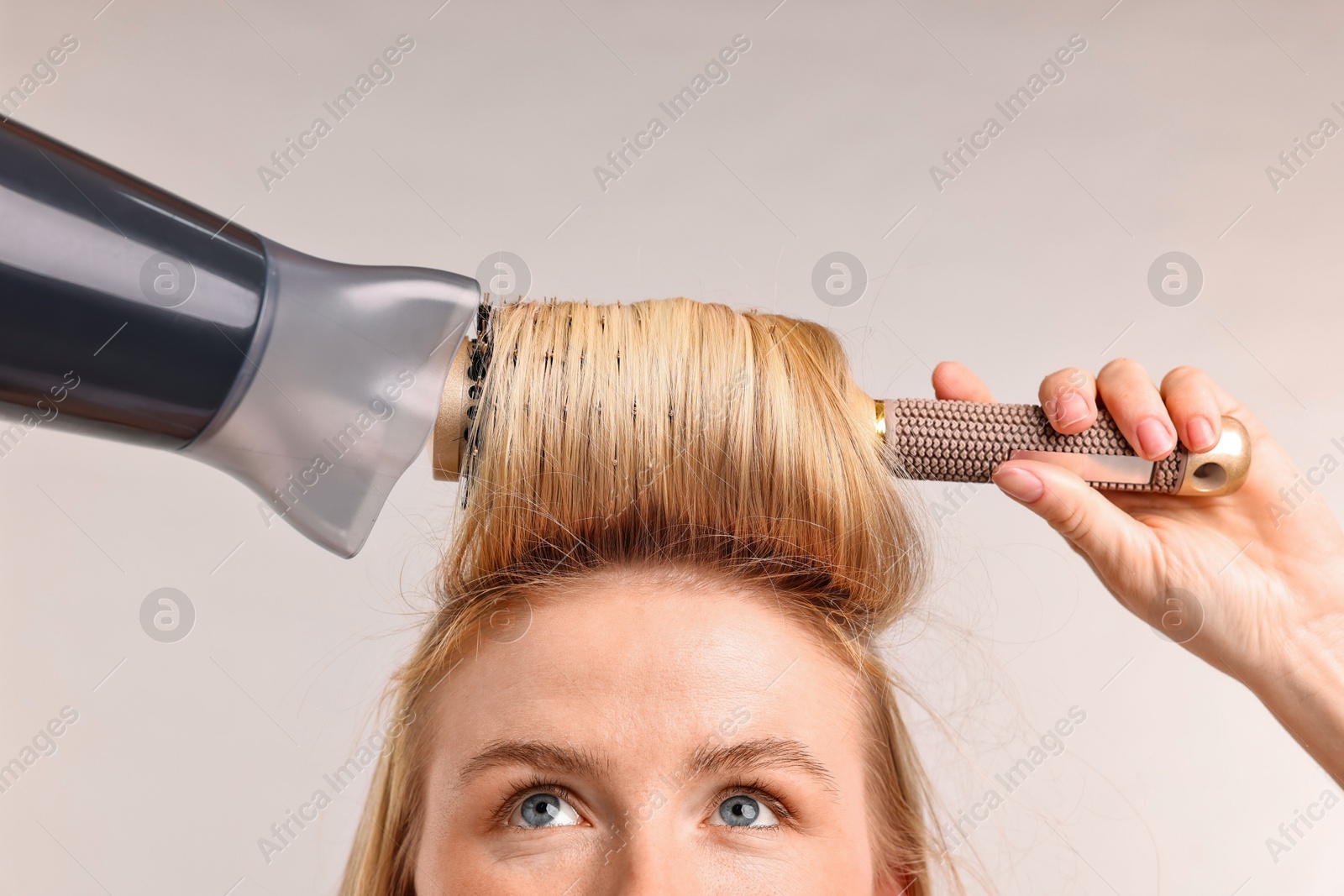 Photo of Beautiful young woman styling her hair with hairdryer and brush on light grey background, closeup