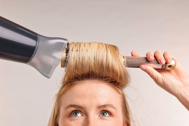 Photo of Beautiful young woman styling her hair with hairdryer and brush on light grey background, closeup