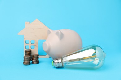 Photo of Piggy bank, coins, house model and lightbulb on light blue background. Energy saving concept