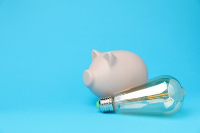 Photo of Piggy bank and lightbulb on light blue background, space for text. Energy saving concept