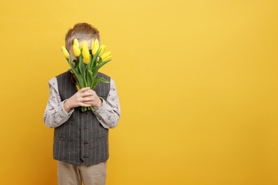 Little boy hiding behind bouquet of tulips on yellow background. Space for text