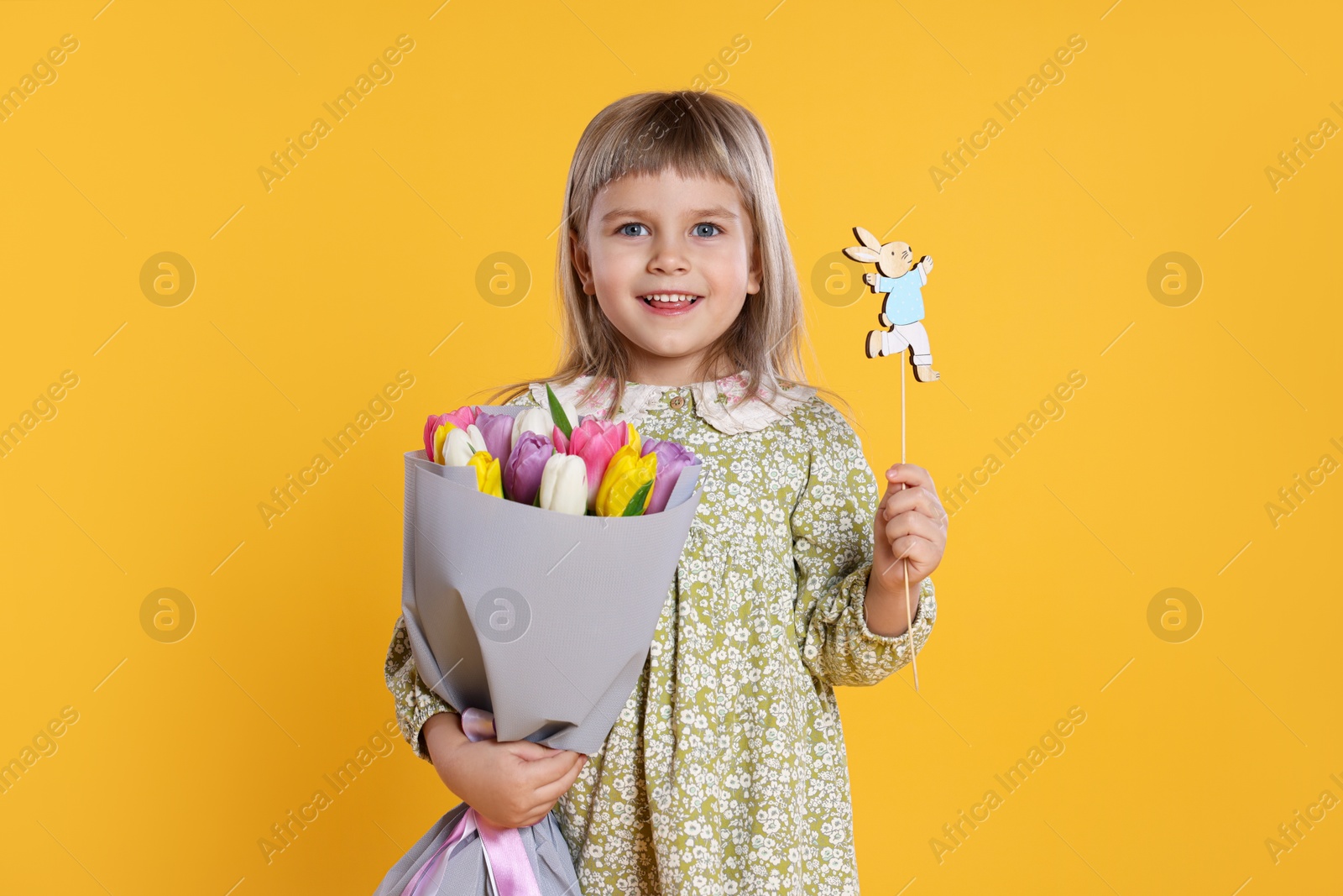 Photo of Smiling little girl with bouquet of tulips and decorative Easter bunny on orange background