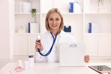 Photo of Weight loss. Smiling nutritionist with stethoscope and scales at table in clinic