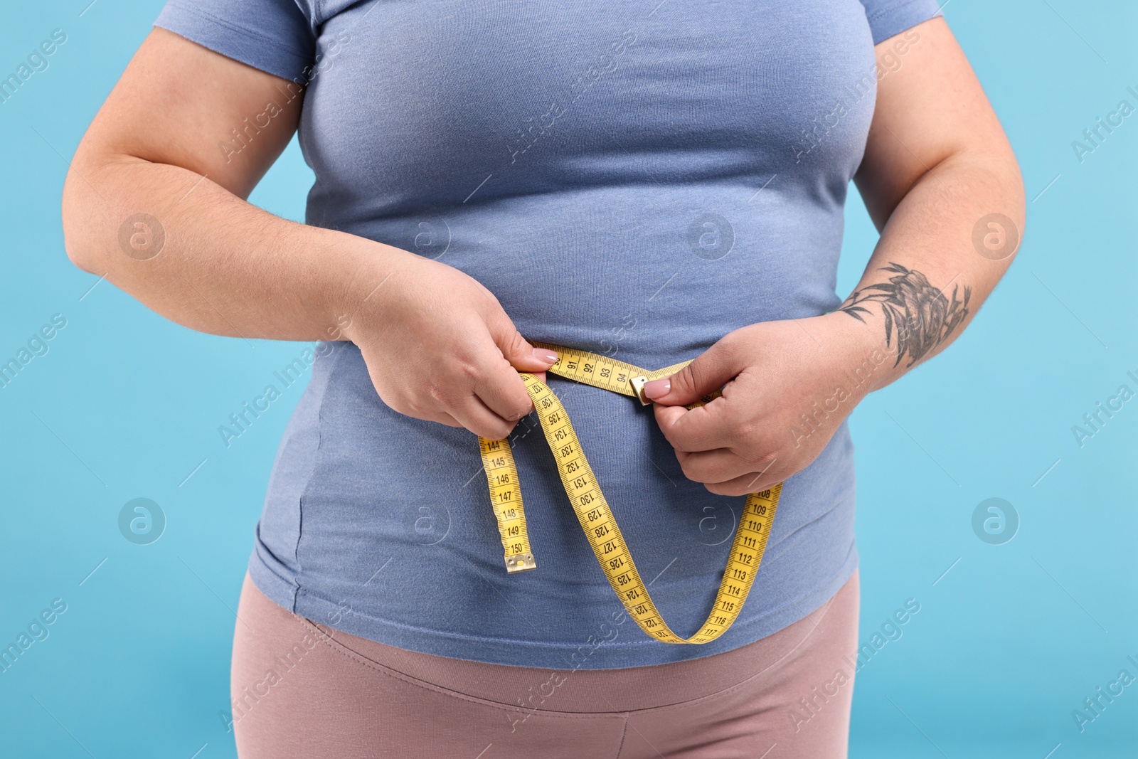 Photo of Weight loss. Plus size woman measuring waist with tape on light blue background, closeup