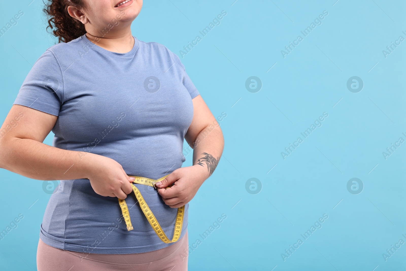 Photo of Weight loss. Happy plus size woman measuring waist with tape on light blue background, closeup with space for text