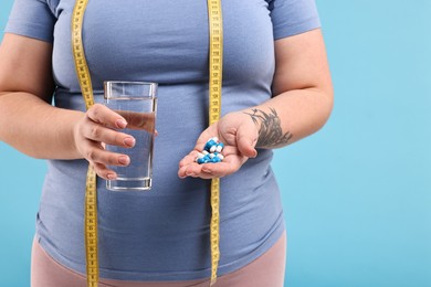 Photo of Plus size woman holding pile of weight loss supplements and glass with water on light blue background, closeup