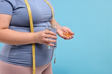 Photo of Plus size woman holding pile of weight loss supplements and glass with water on light blue background, closeup. Space for text