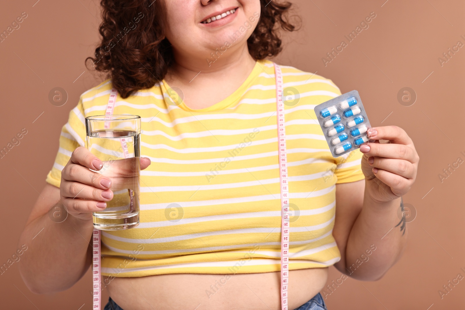 Photo of Happy plus size woman holding blister of weight loss supplements and glass of water on beige background, closeup