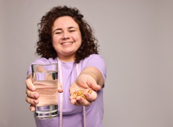 Photo of Happy plus size woman giving pile of weight loss supplements and glass of water on grey background, selective focus. Space for text