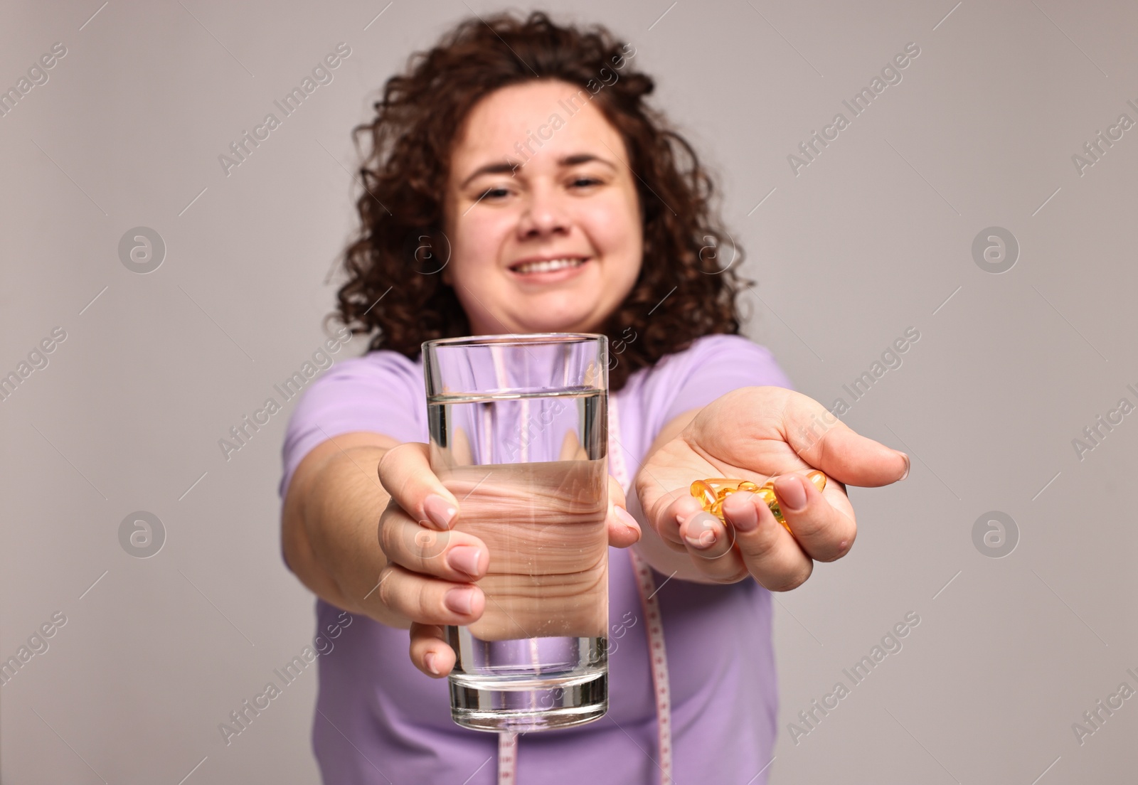 Photo of Happy plus size woman giving pile of weight loss supplements and glass of water on grey background, selective focus