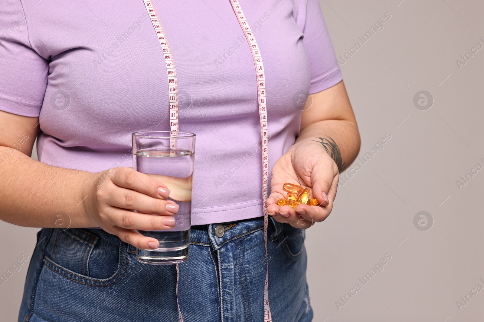 Photo of Plus size woman with pile of weight loss supplements and glass of water on grey background, closeup