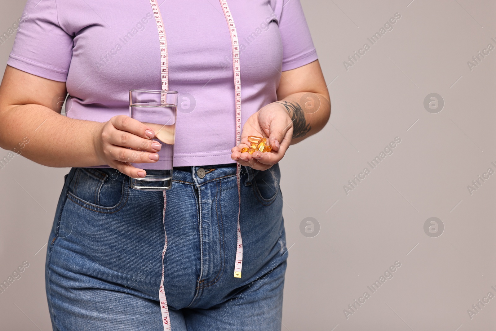 Photo of Plus size woman with pile of weight loss supplements and glass of water on grey background, closeup. Space for text