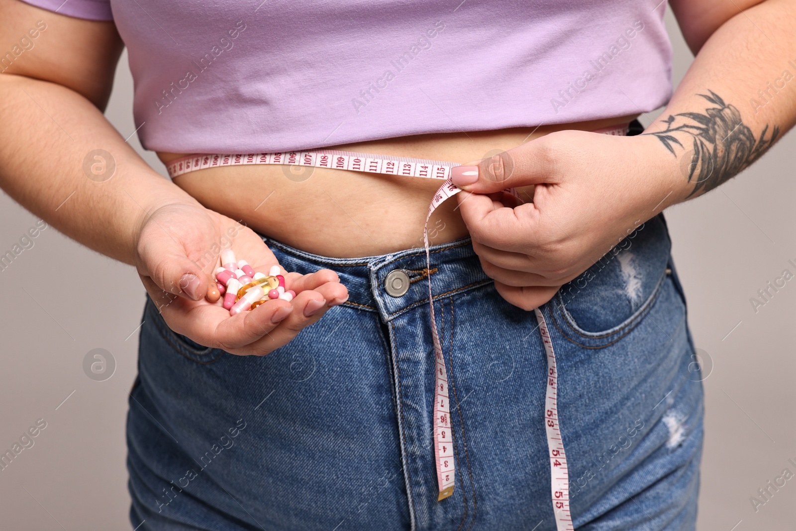 Photo of Plus size woman measuring waist with tape and holding pile of weight loss supplements on grey background, closeup