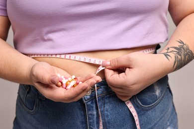 Photo of Plus size woman measuring waist with tape and holding pile of weight loss supplements on grey background, closeup