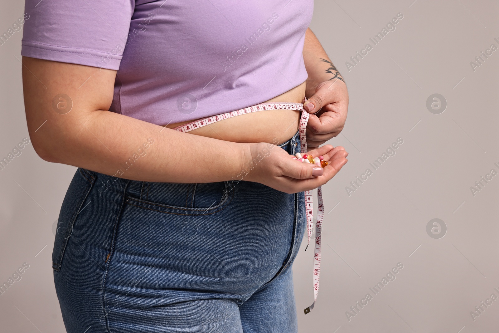 Photo of Plus size woman measuring waist with tape and holding pile of weight loss supplements on grey background, closeup. Space for text