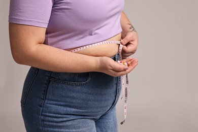 Photo of Plus size woman measuring waist with tape and holding pile of weight loss supplements on grey background, closeup. Space for text