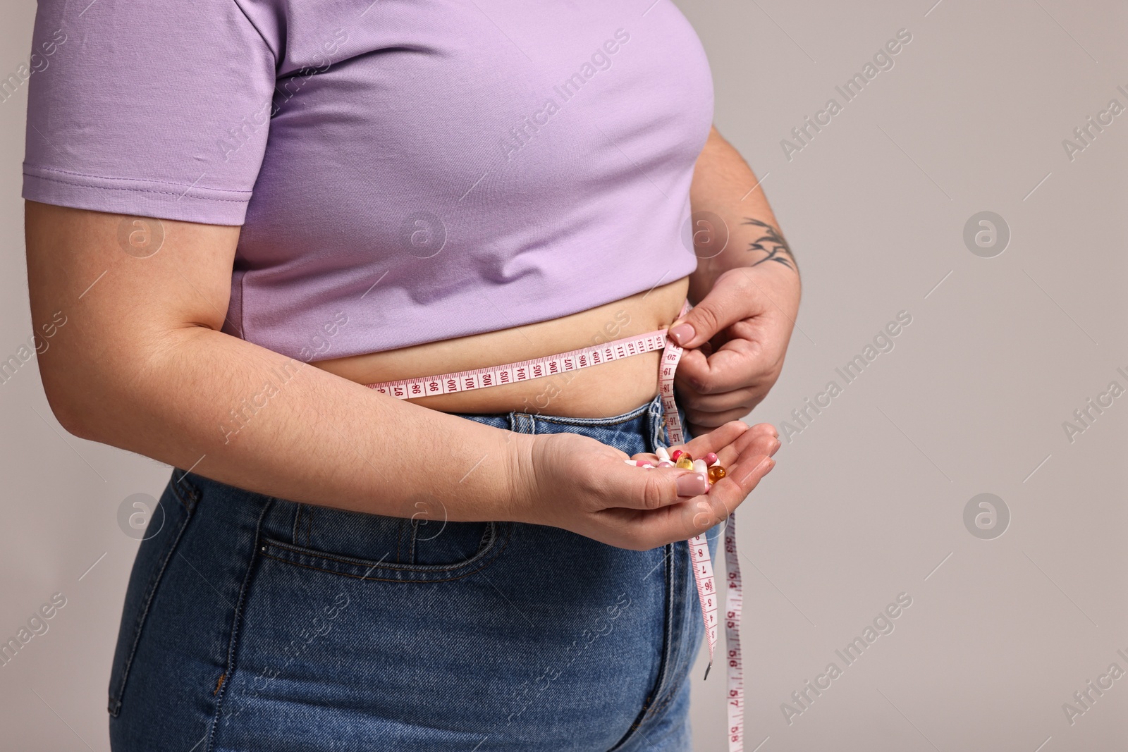 Photo of Plus size woman measuring waist with tape and holding pile of weight loss supplements on grey background, closeup