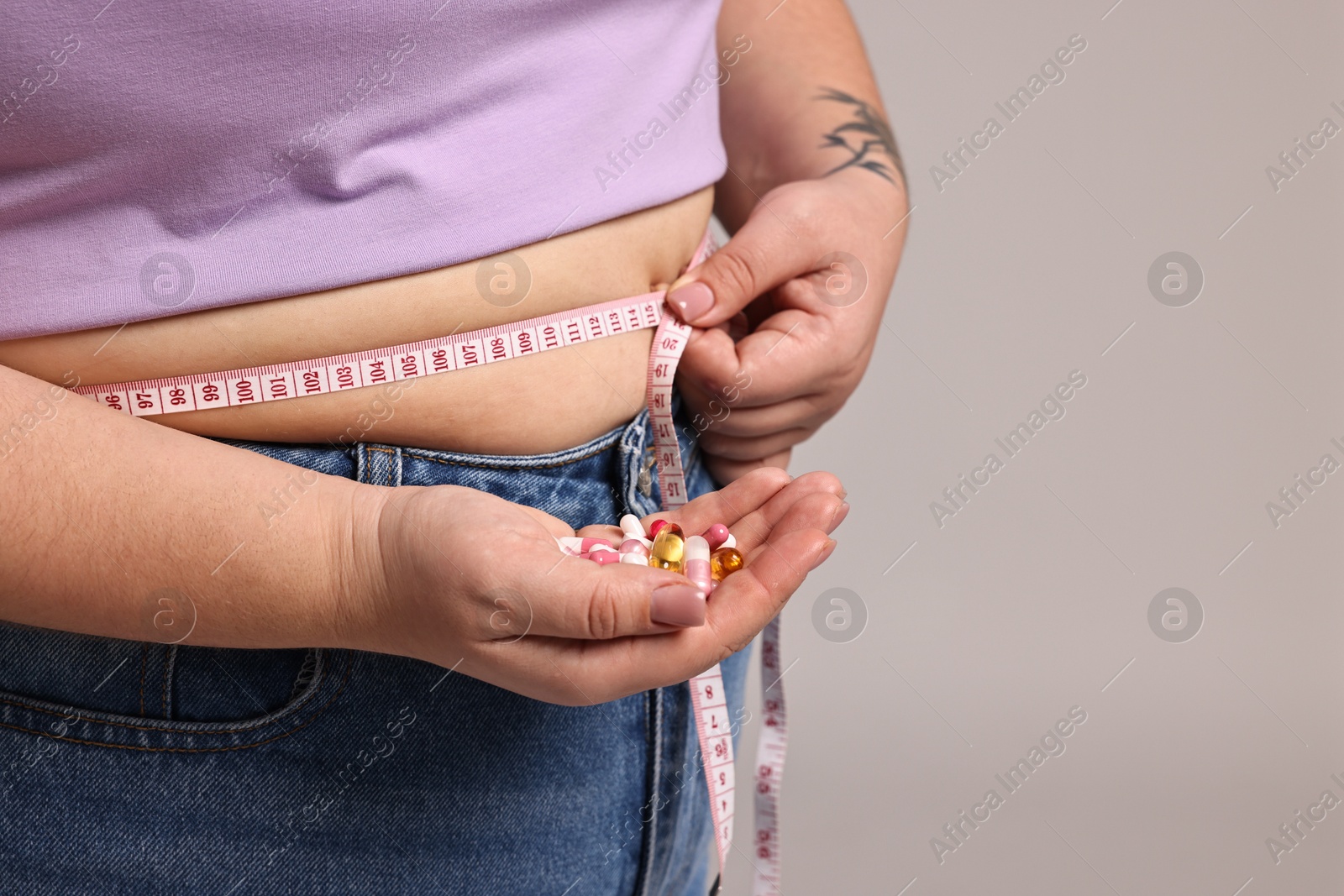 Photo of Plus size woman measuring waist with tape and holding pile of weight loss supplements on grey background, closeup. Space for text