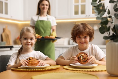 Photo of Cute kids eating tasty buns at table indoors, selective focus. Woman holding baking pan with pastry in kitchen