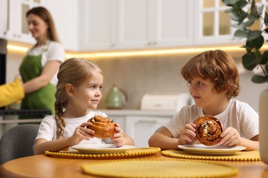 Photo of Cute kids eating tasty buns at table while their mother baking in kitchen, selective focus