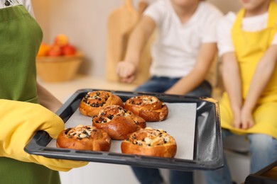 Photo of Mother holding baking pan with tasty buns near her kids in kitchen, closeup