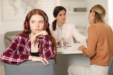 Photo of Annoyed teenage girl listening to music instead of working with psychologist in office, selective focus. Mother and daughter difficult relationship