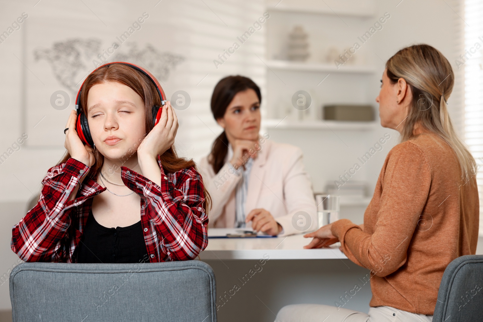 Photo of Annoyed teenage girl listening to music instead of working with psychologist in office, selective focus. Mother and daughter difficult relationship