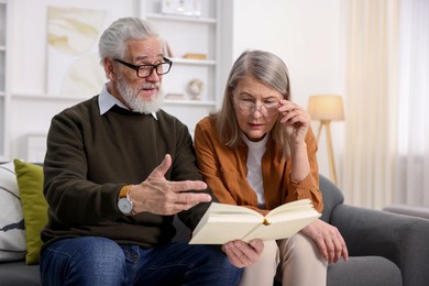 Photo of Cute elderly couple reading book together on sofa at home