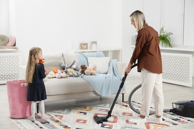 Photo of Little helper. Girl putting away toys while her mother vacuuming at home