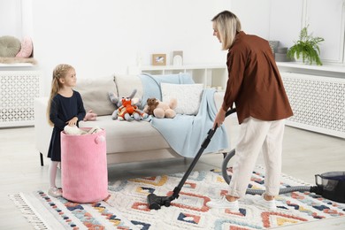 Photo of Little helper. Girl putting away toys while her mother vacuuming at home