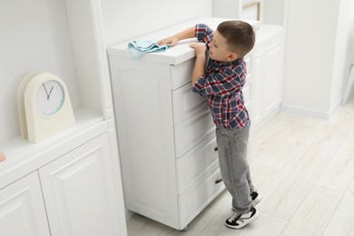 Photo of Little helper. Cute boy wiping dust from chest of drawers at home